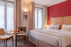 A hotel room with a bed, a table with breakfast items, a chair, and a window with curtains.