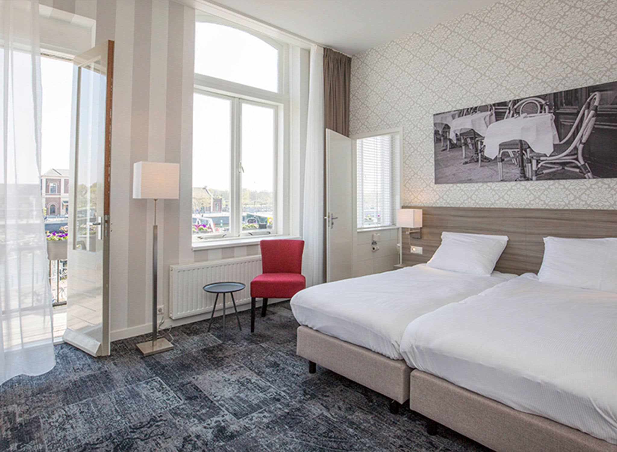 A hotel room with a large bed, a red chair, a small table, and a window with a view of the street.