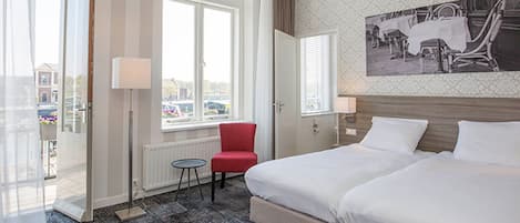 A hotel room with a large bed, a red chair, a small table, and a window with a view of the street.