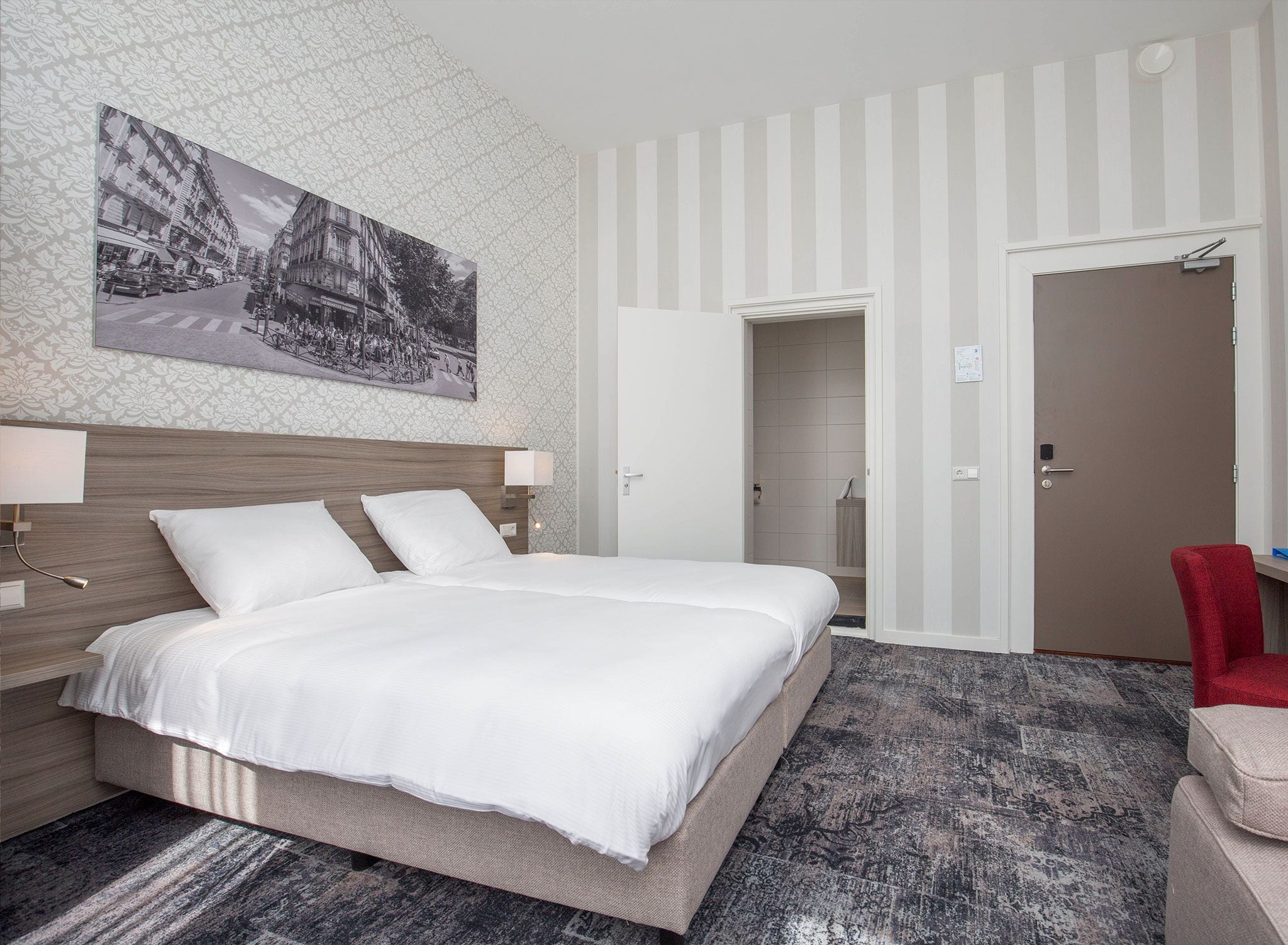 Una habitación de hotel con una cama grande, cabecera, mesita de noche, una silla roja y una puerta que da al baño.