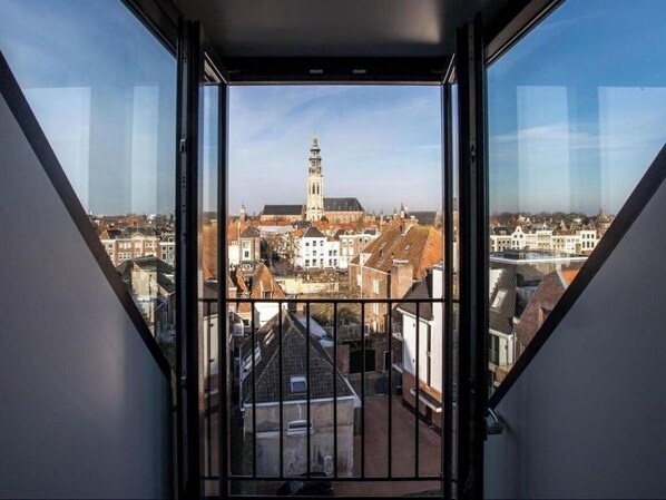 A view from a window with a metal railing, overlooking a town with historic buildings and a prominent tower.