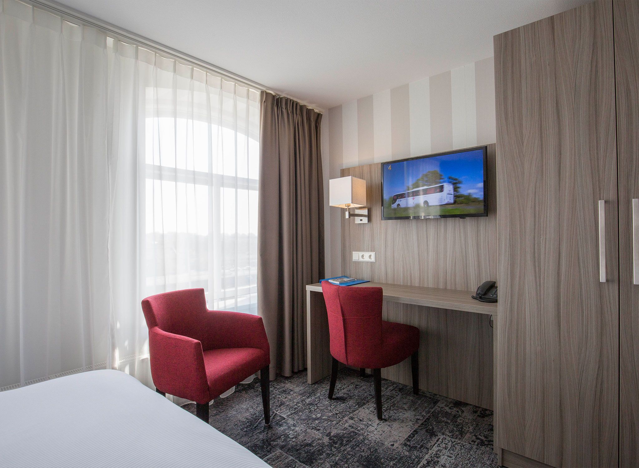 Une chambre d’hôtel avec un lit, deux fauteuils rouges, un bureau avec une lampe, une télévision et une grande fenêtre avec des rideaux.