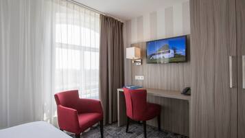 Une chambre d’hôtel avec un lit, deux fauteuils rouges, un bureau avec une lampe, une télévision et une grande fenêtre avec des rideaux.