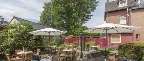 An outdoor patio area with wicker tables and chairs, umbrellas, and a brick building in the background.