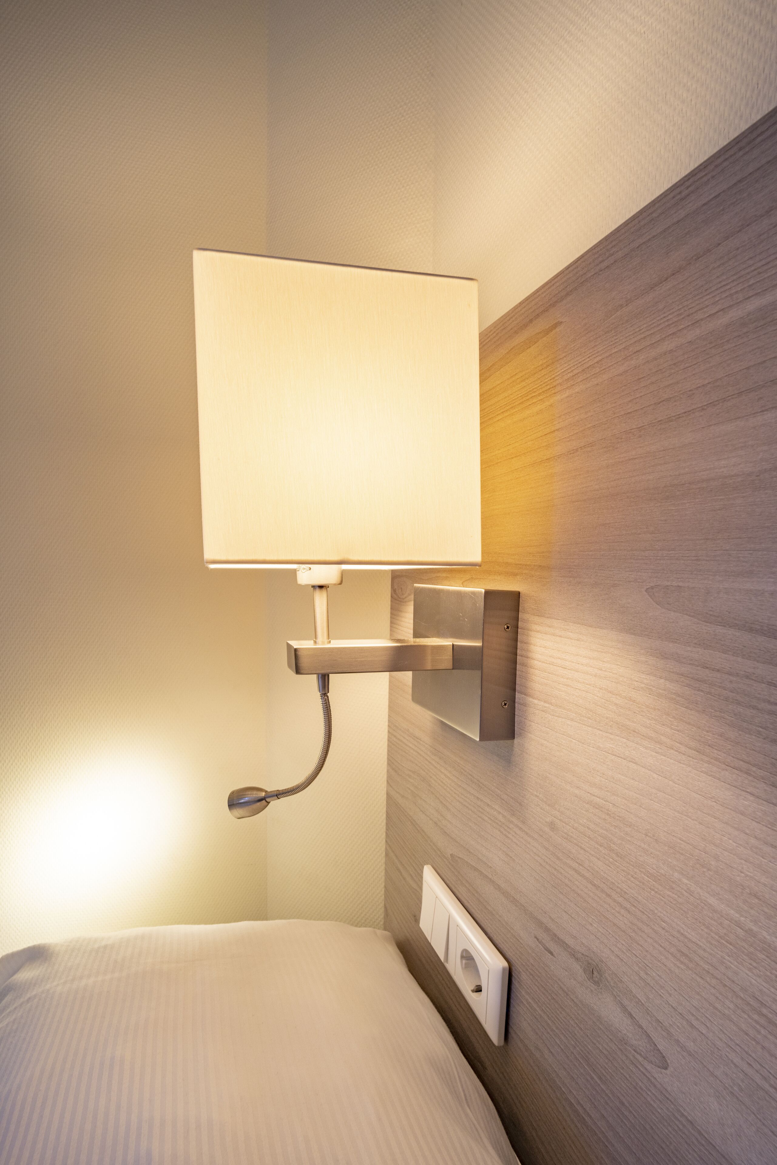 A modern hotel room with a wall-mounted lamp, a bedside table, and a power outlet.