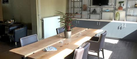 A modern dining area with a wooden table and chairs, a potted plant, and a kitchen area with a TV and shelving.