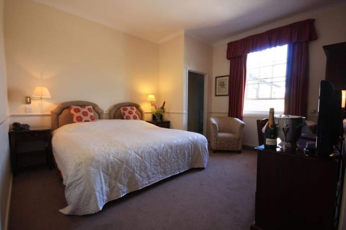 A hotel room with a large bed, two bedside tables with lamps, a chair, a desk with a computer and a bottle of champagne, and a window with curtains.