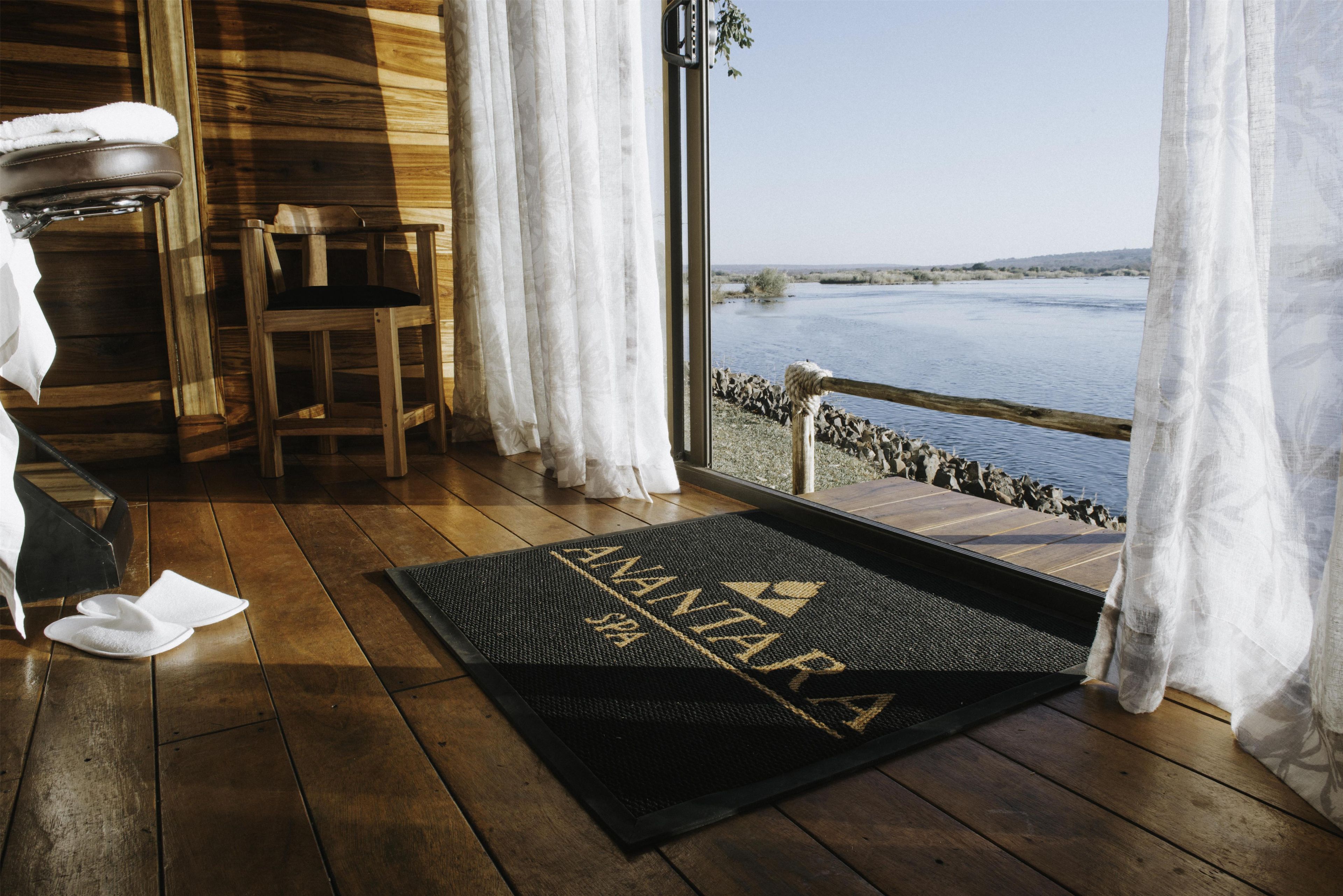 Uma cabana de madeira com soalho em madeira, um banco, uma cadeira, um tapete na porta e vista para um corpo d'água através de uma janela com cortinas.