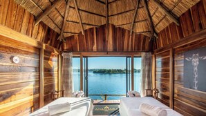 A wooden cabin with two beds, a thatched roof, and a view of a lake. - Royal Livingstone Victoria Falls Zambia Hotel by Anantara (Livingstone)