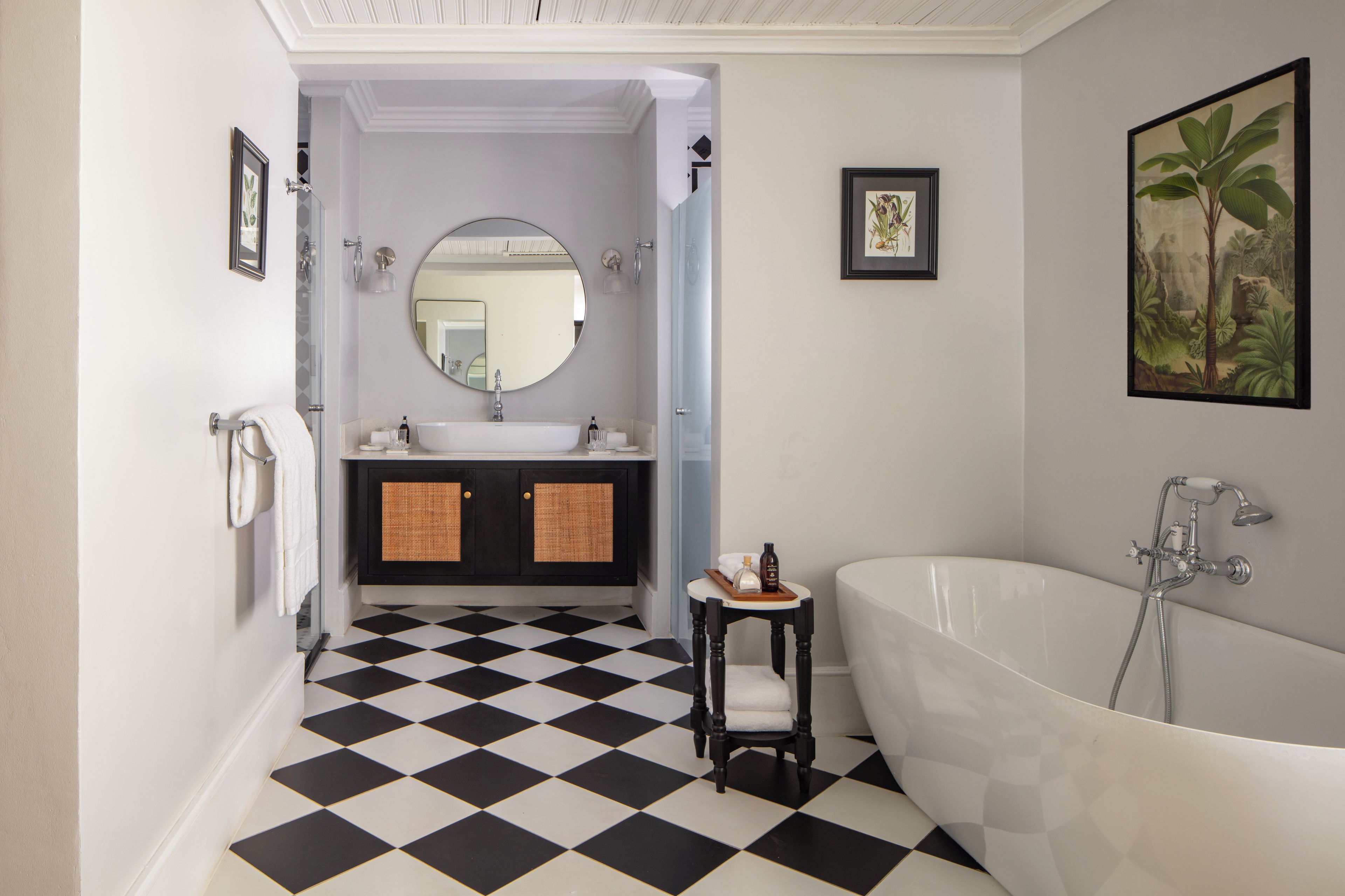 Ein Badezimmer mit Schachbrettmuster-Boden, einer freistehenden Badewanne, einem runden Spiegel und einem gerahmten Pflanzenmotiv an der Wand.