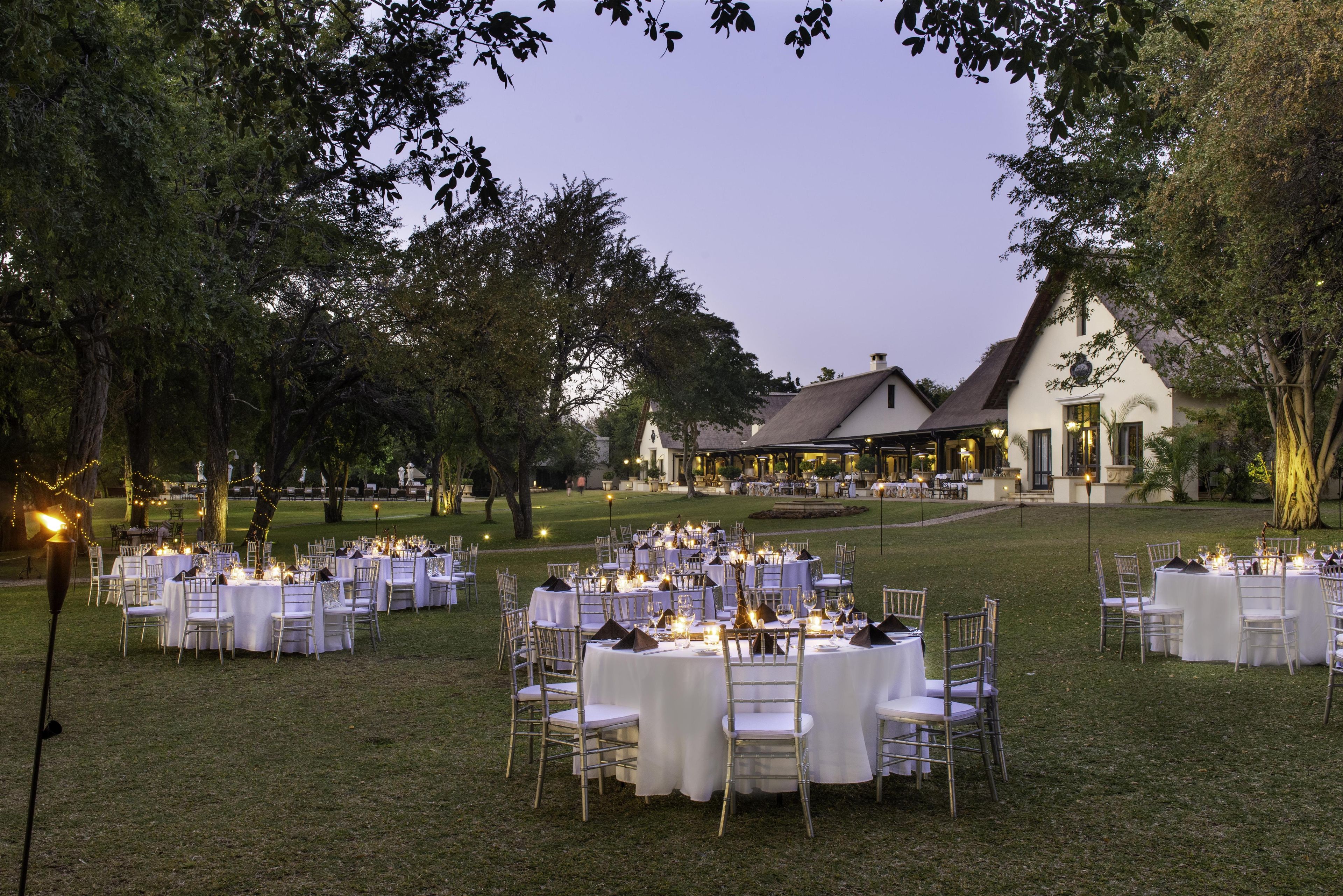 An outdoor banquet setup with round tables and chairs, a building in the background, and trees surrounding the area.