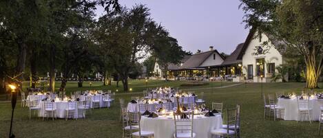 An outdoor banquet setup with round tables and chairs, a building in the background, and trees surrounding the area.