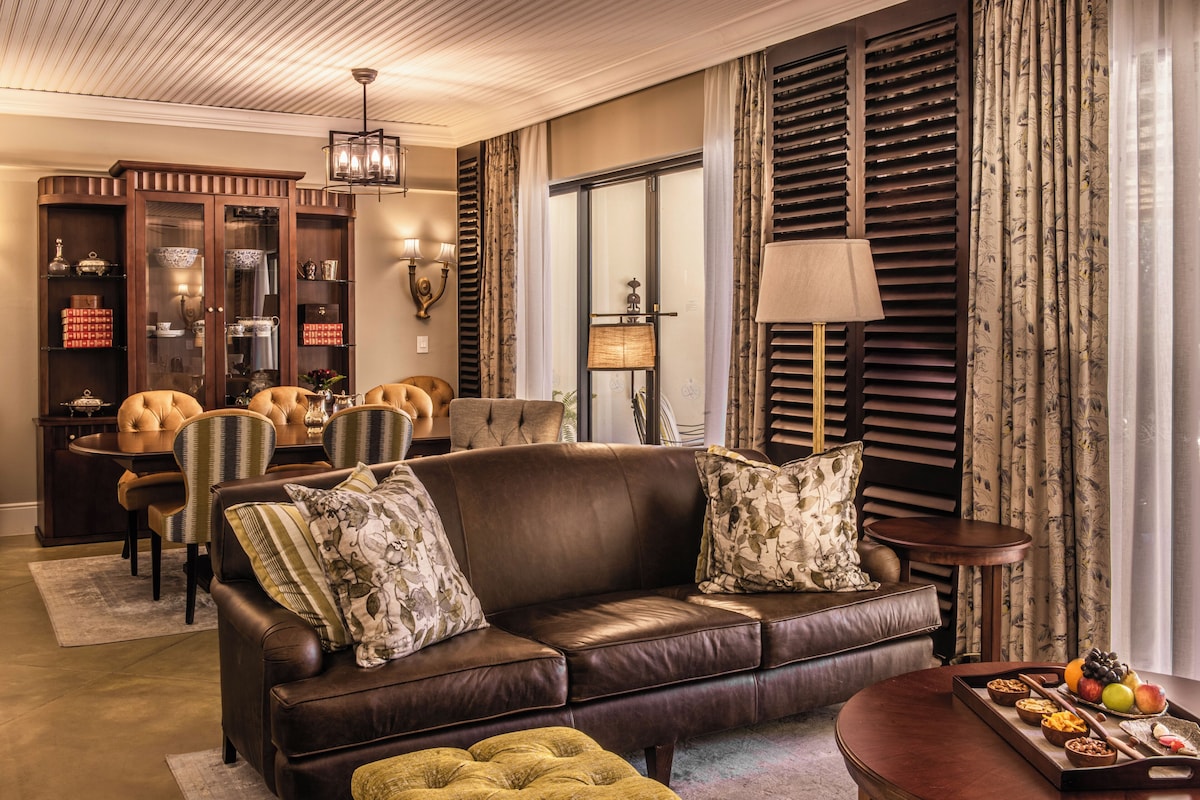 A living room with a brown leather sofa, a dining table with chairs, a wooden cabinet with glass doors, and a lamp on a side table.