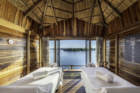 A wooden cabin with two beds, a view of a lake, and a thatched roof. - Royal Livingstone Victoria Falls Zambia Hotel by Anantara (Livingstone)