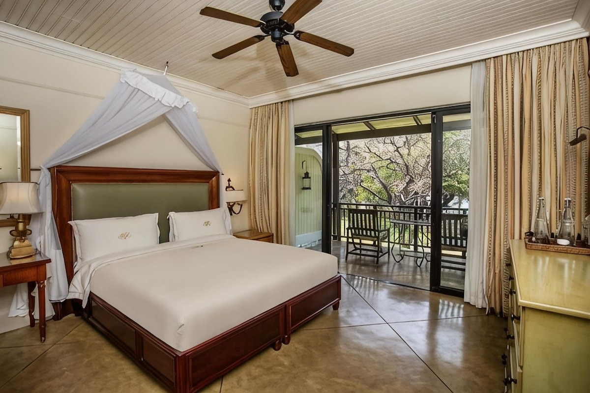 A hotel room with a four-poster bed, a ceiling fan, a balcony with a view of trees, and a wooden nightstand with a lamp.
