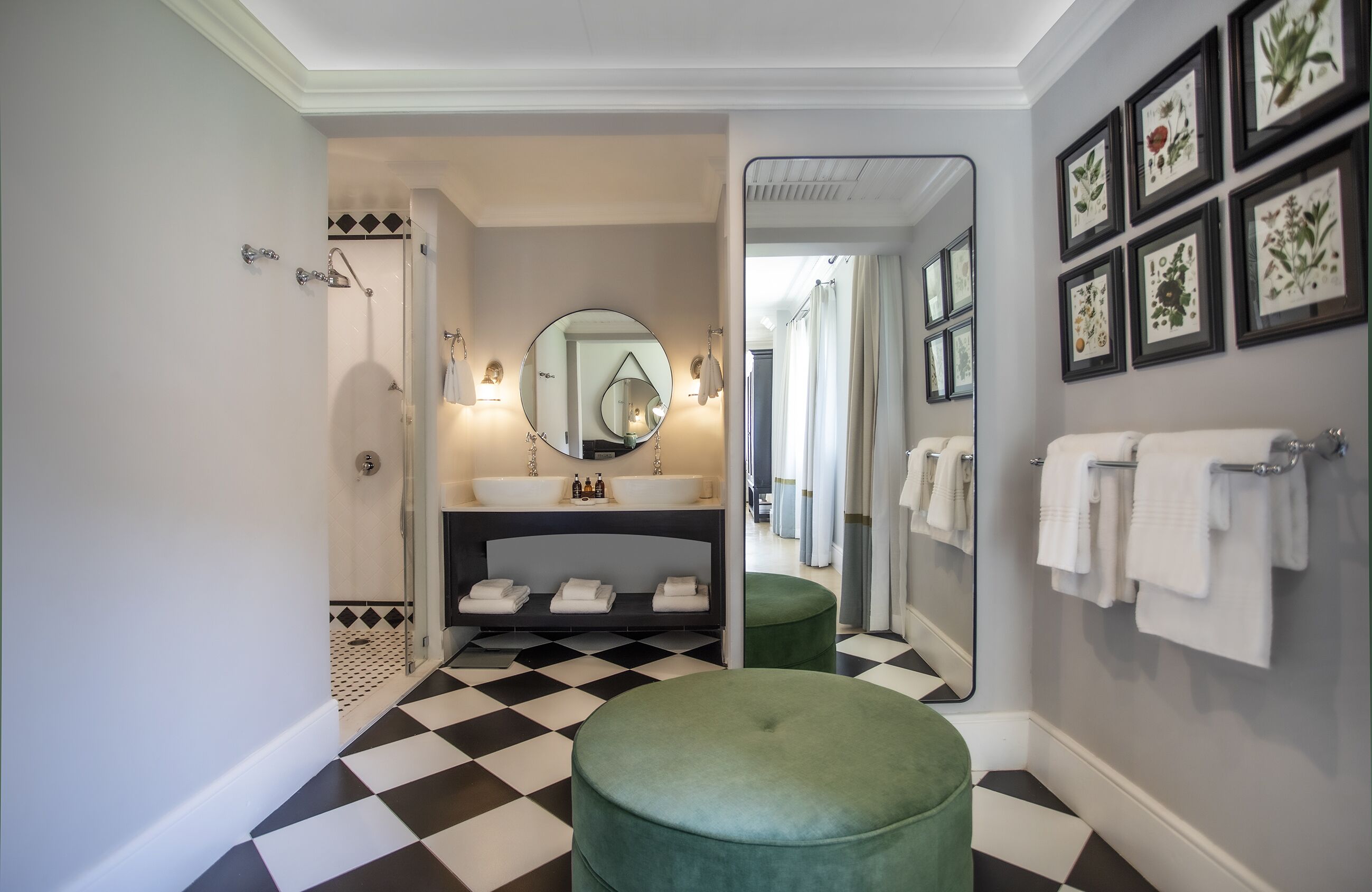 A bathroom with a checkered floor, a green ottoman, a sink with two mirrors, and framed botanical prints on the wall.