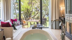 A room with a large window, a bathtub, a sofa with pillows, a table with a hat and a bottle, and a cabinet with a mirror. - Royal Livingstone Victoria Falls Zambia Hotel by Anantara (Livingstone)