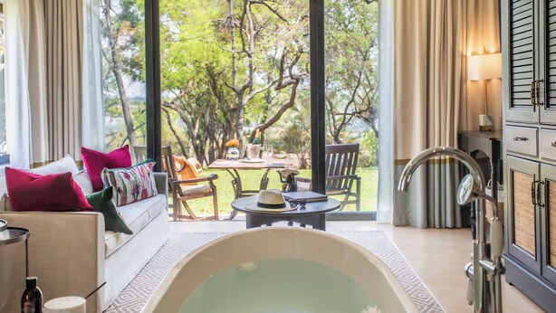 A room with a large window, a bathtub, a sofa with pillows, a table with a hat and a bottle, and a cabinet with a mirror.