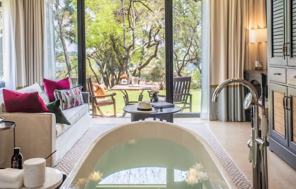 A room with a large window, a bathtub, a sofa with pillows, a table with a hat and a bottle, and a cabinet with a mirror.