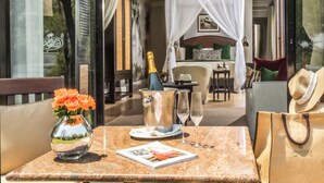 A sunlit outdoor seating area with a table set for two, featuring a bottle of champagne, a vase of flowers, and a magazine.