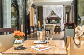 A sunlit outdoor seating area with a table set for two, featuring a bottle of champagne, a vase of flowers, and a magazine.