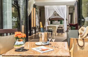 Een zonnig buitengebied met een tafel voor twee, compleet met een champagnefles, een bloevas en een tijdschrift.