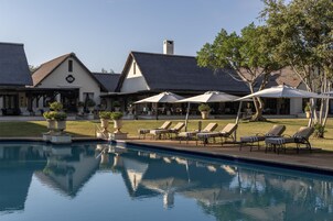 Zona de piscina con sillones de descanso y sombrillas, rodeada por un edificio con techo de paja.