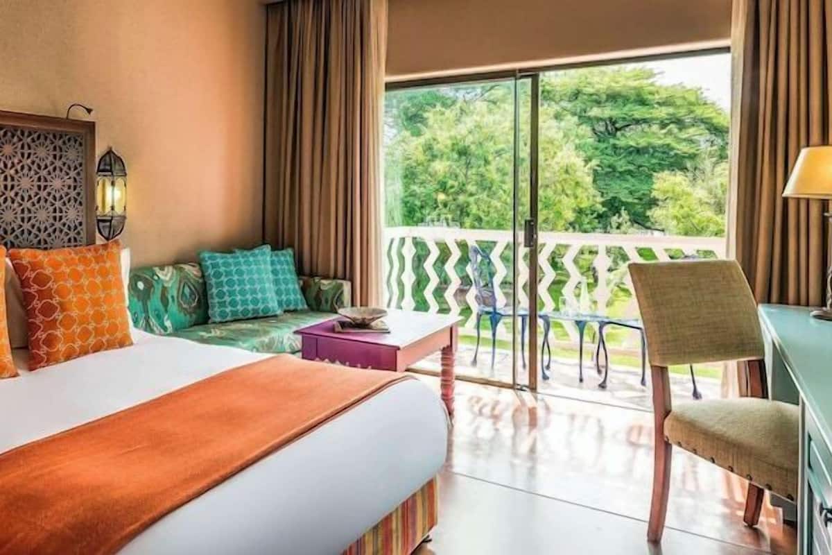 A hotel room with a large bed, a desk, a chair, a balcony with a view of greenery, and a decorative headboard.