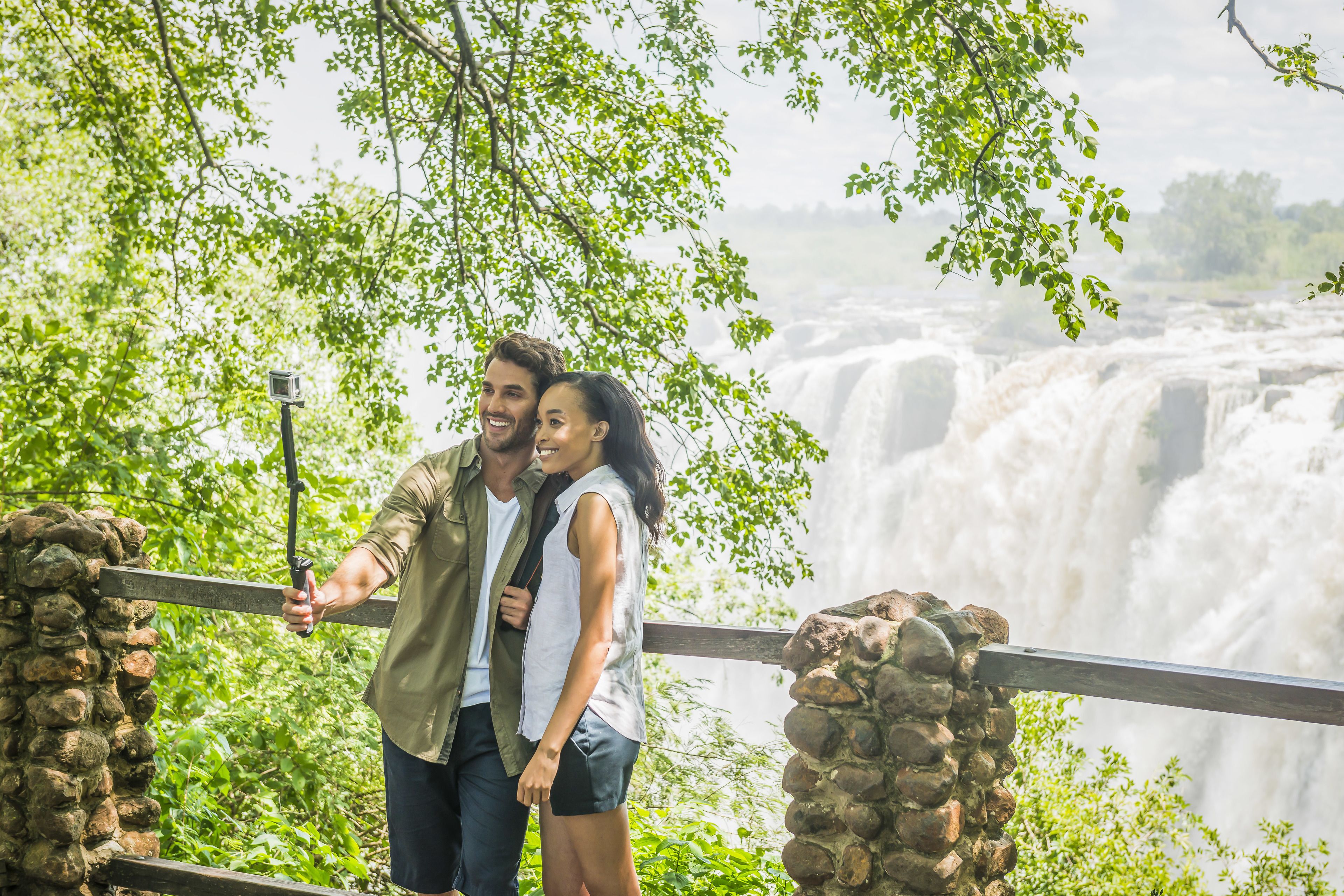 A couple standing by a railing overlooking a waterfall, with stone pillars and greenery in the background.