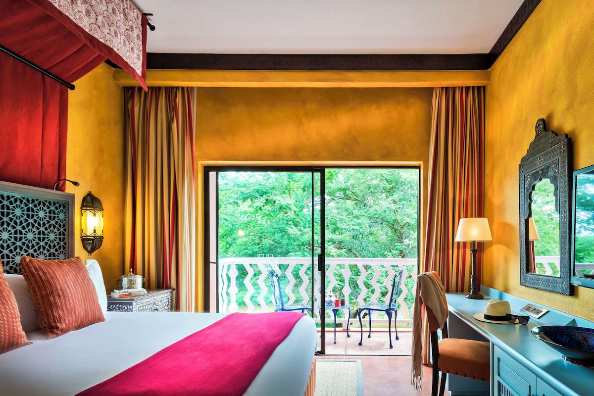 A hotel room with a bed, a desk, a chair, a balcony with a railing, and a view of greenery outside.