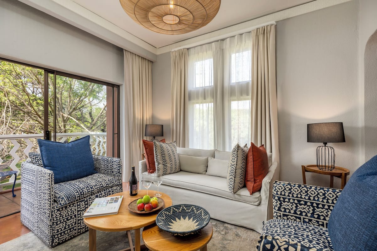 A modern living room with patterned furniture, a dining table with fruit, and a view of trees through large windows.
