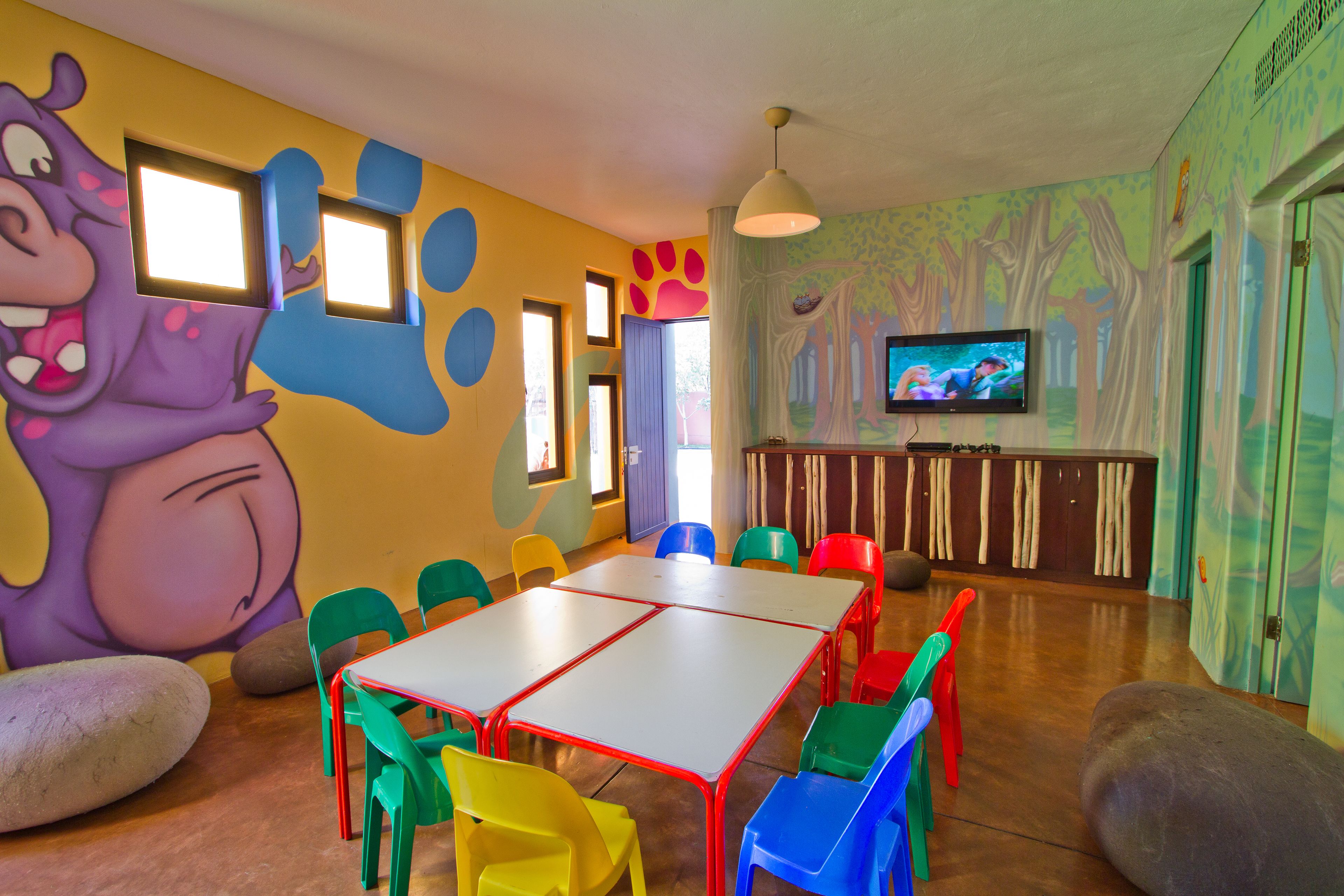 A children's playroom with colorful walls, a large cartoon character mural, a TV, and a table with chairs.