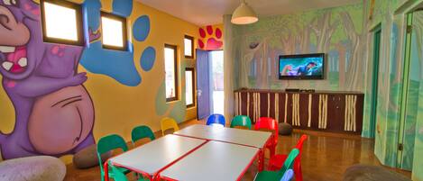 A children's playroom with colorful walls, a large cartoon character mural, a TV, and a table with chairs.