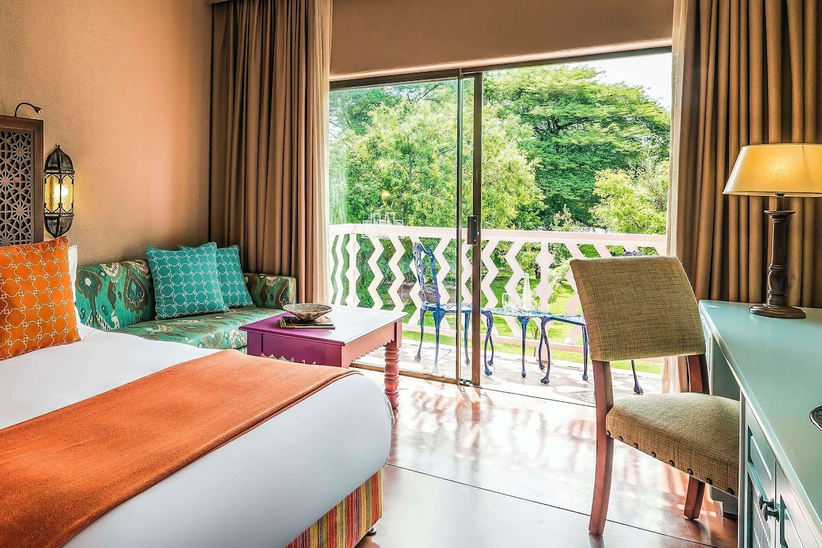 A hotel room with a large bed, a balcony with a view of trees, a desk with a lamp, and a decorative wall panel.