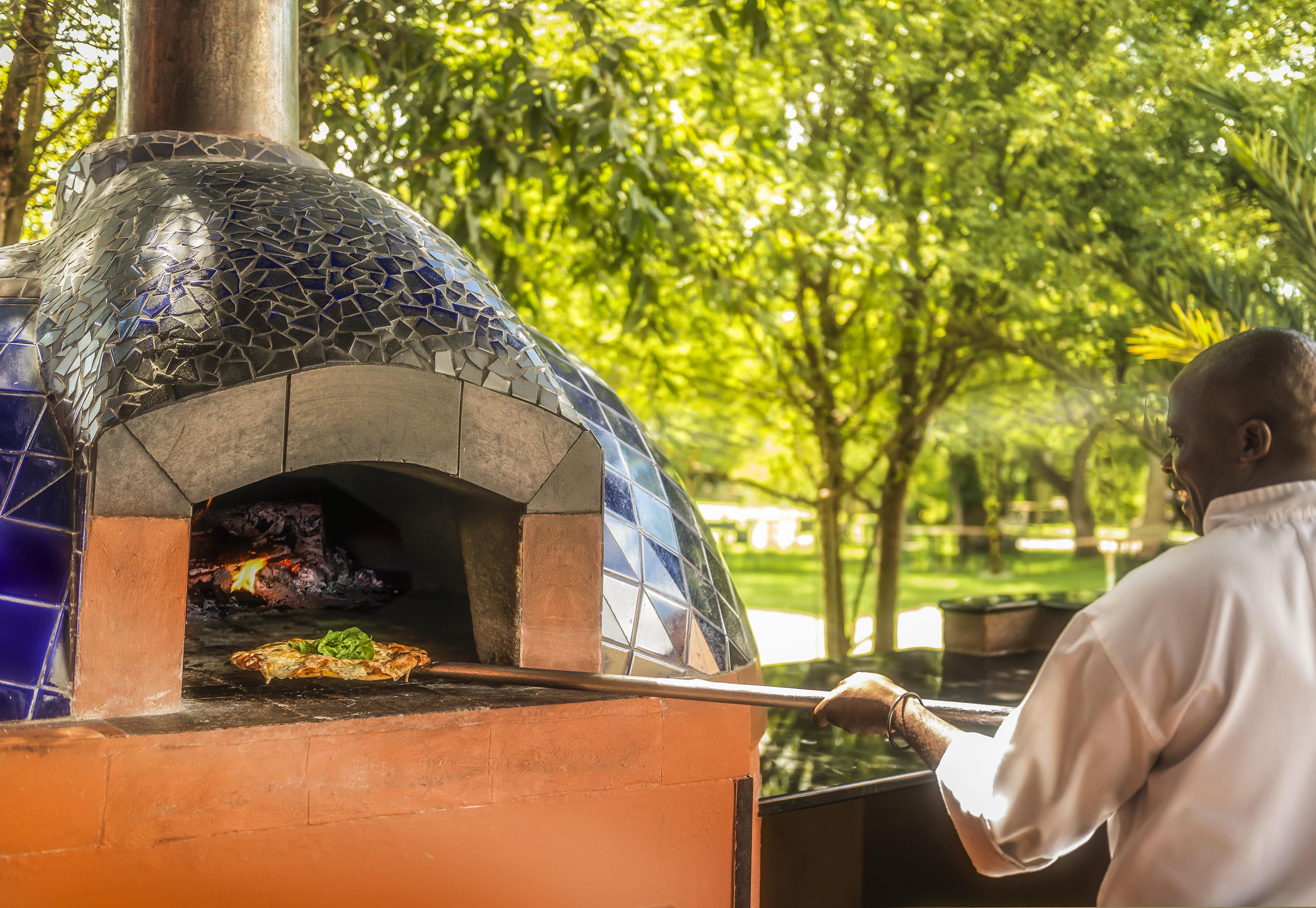 An outdoor brick oven pizza oven with a person using a long-handled tool to retrieve a pizza.