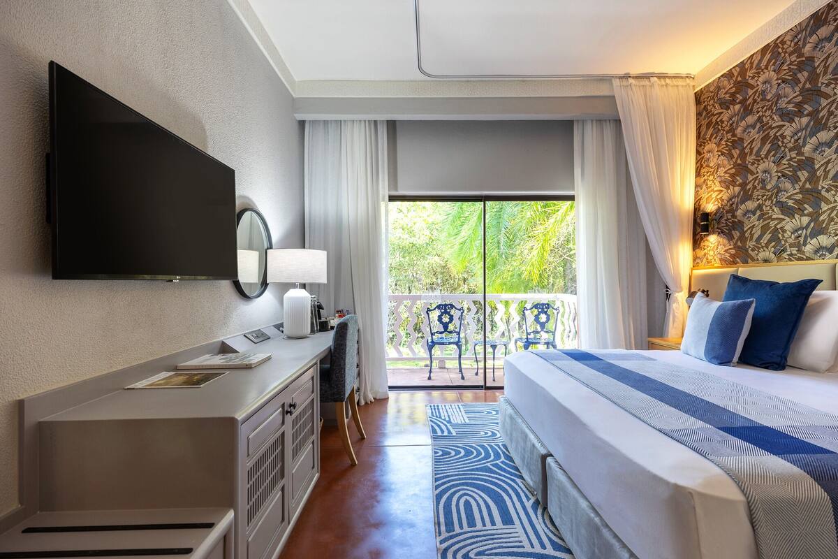 A hotel room with a flat-screen TV, a desk with a lamp, a bed with blue and white bedding, a patterned rug, and a balcony with chairs and curtains.