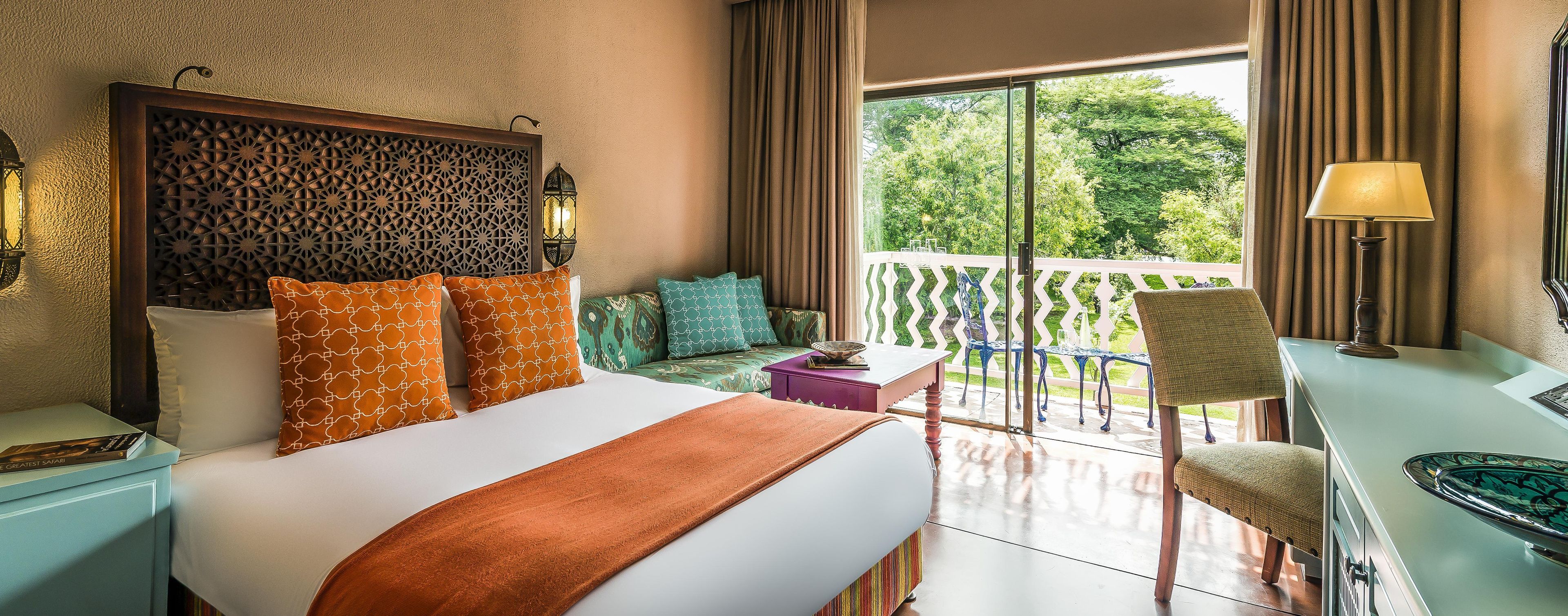 Habitación de hotel con cama, escritorio, silla, balcón con barandilla y vista a árboles.