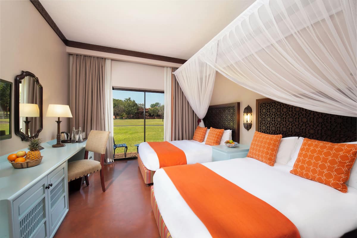 A hotel room with two beds, a desk with a lamp, a chair, and a view of the outdoors.