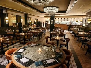 A restaurant with round tables, chairs, chandeliers, and a bar area.