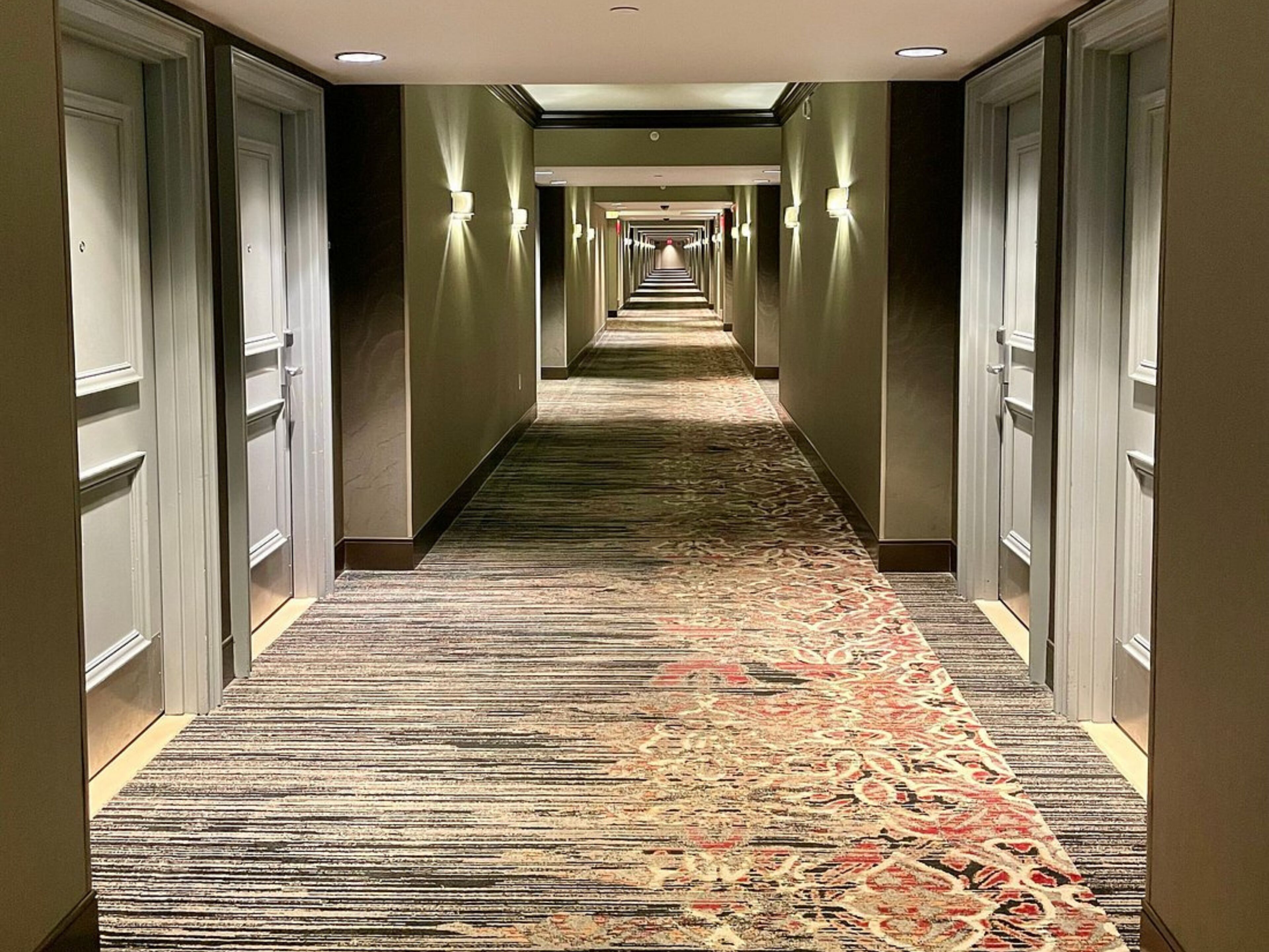 A hotel hallway with carpeted floors, doors on both sides, and wall-mounted lighting.