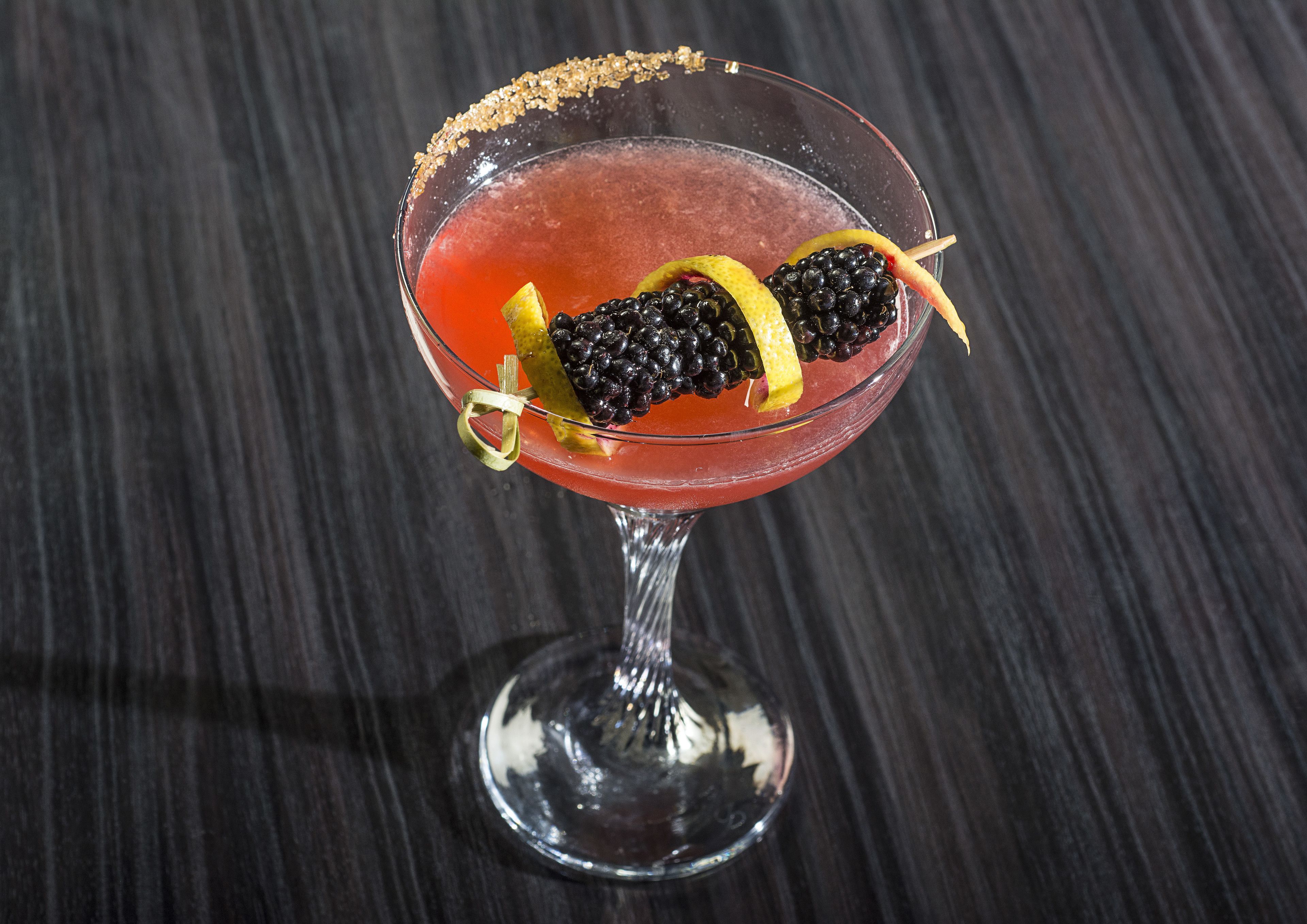 A cocktail in a stemmed glass with a sugar rim, garnished with blackberries and a lemon slice.