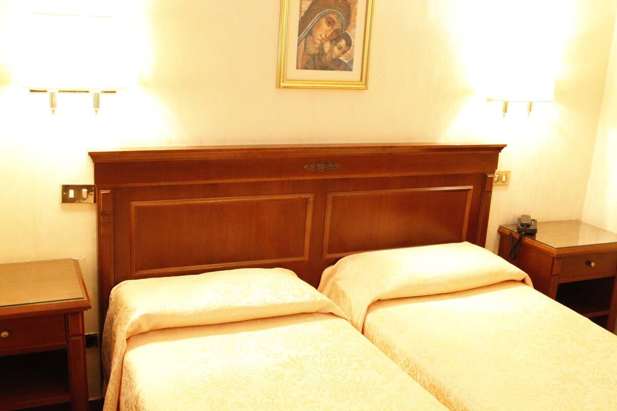 A hotel room with a wooden headboard, two beds with matching linens, bedside tables, and a framed picture on the wall.