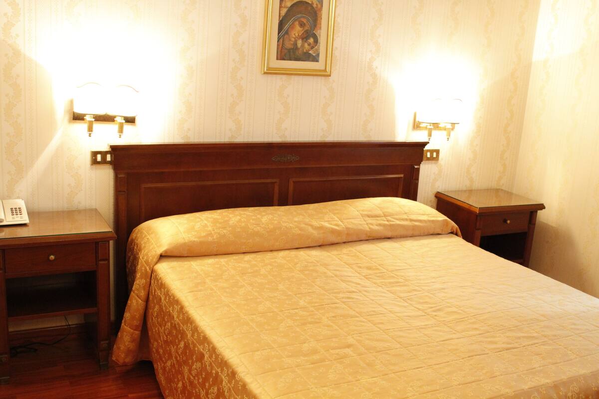 A hotel room with a wooden headboard, a bed with a patterned bedspread, two bedside tables with lamps, and a wall-mounted telephone.