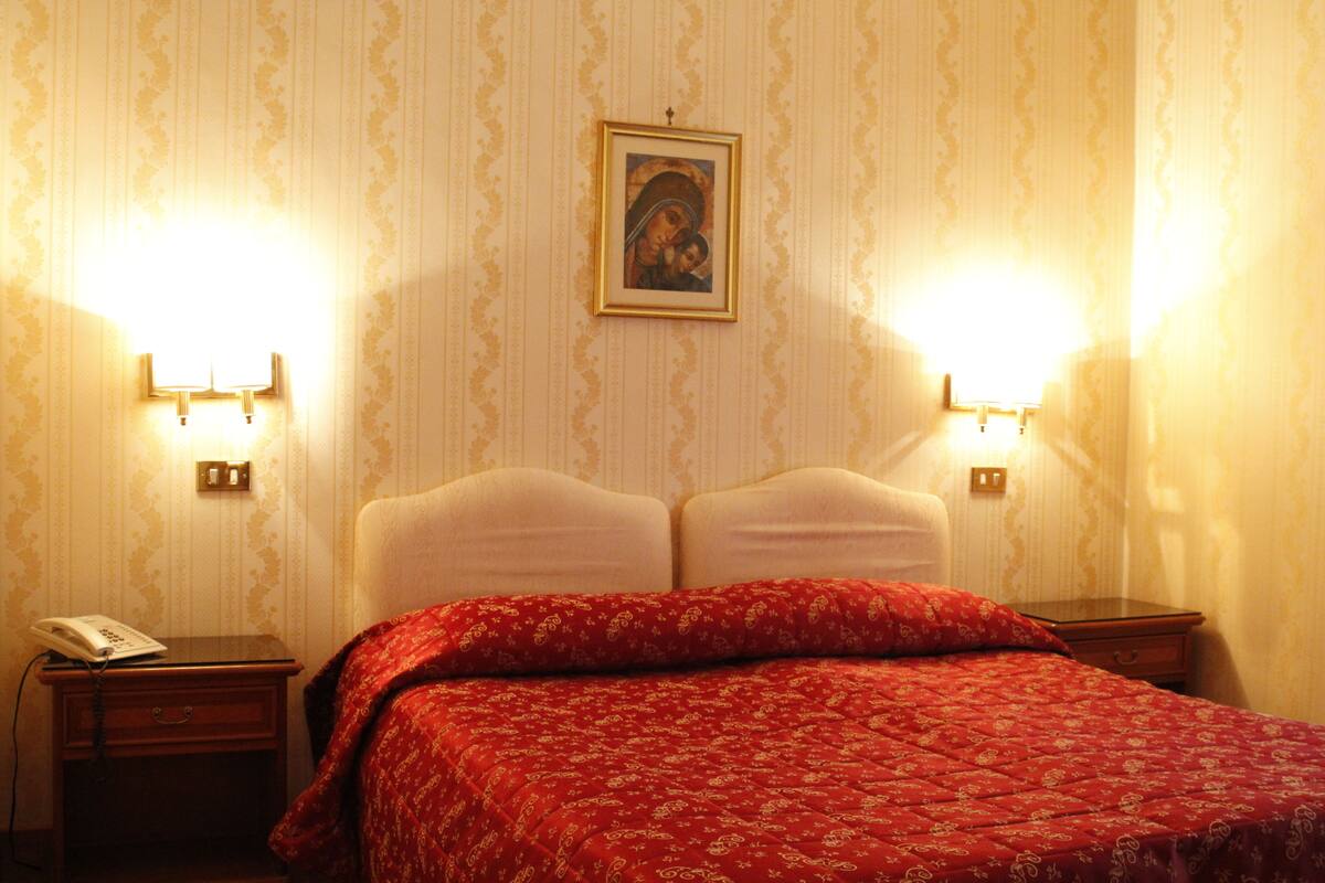 A hotel room with a bed, two bedside tables, a nightstand, a framed picture on the wall, and two wall-mounted lamps.