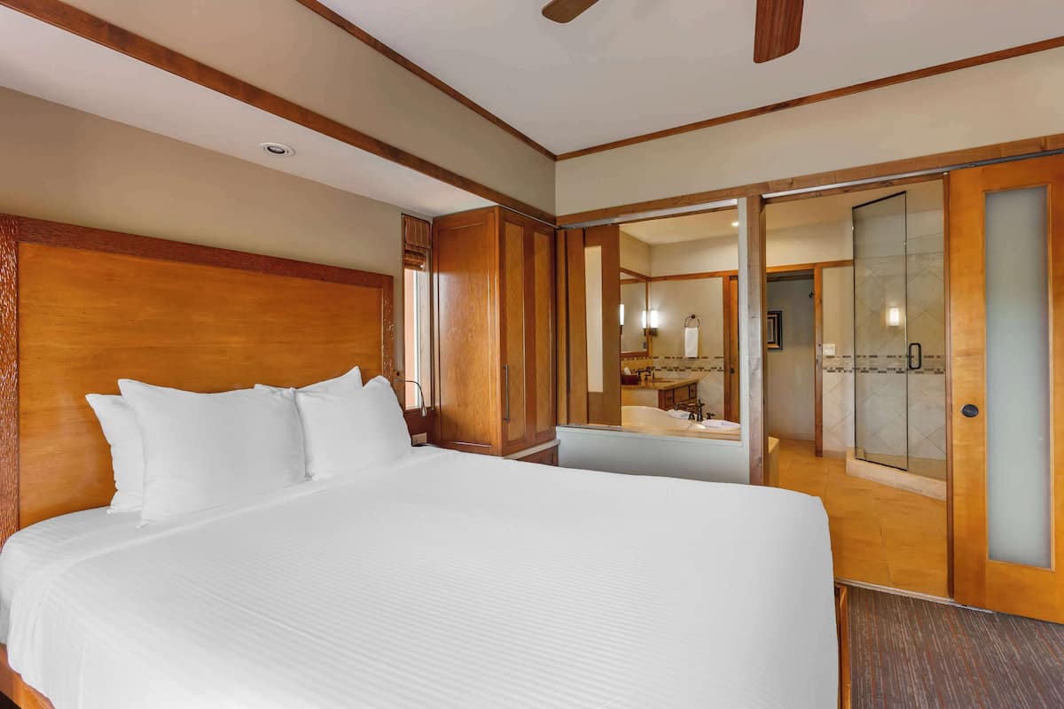 Quarto de hotel com uma cama grande, cabeceira de madeira, banheiro com chuveiro de vidro e piso de madeira.