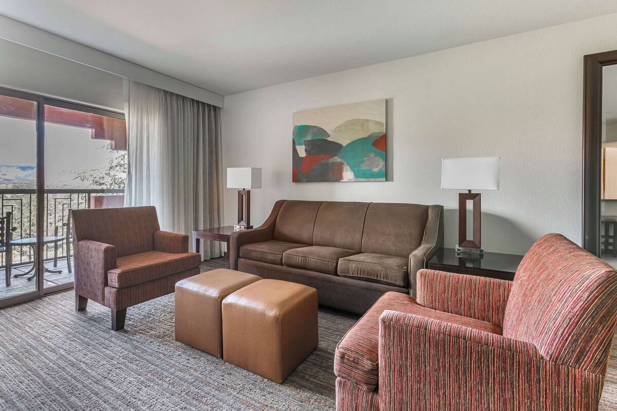 Quarto de hotel com uma janela grande, um sofá, uma poltrona, uma mesa e um quadro na parede.