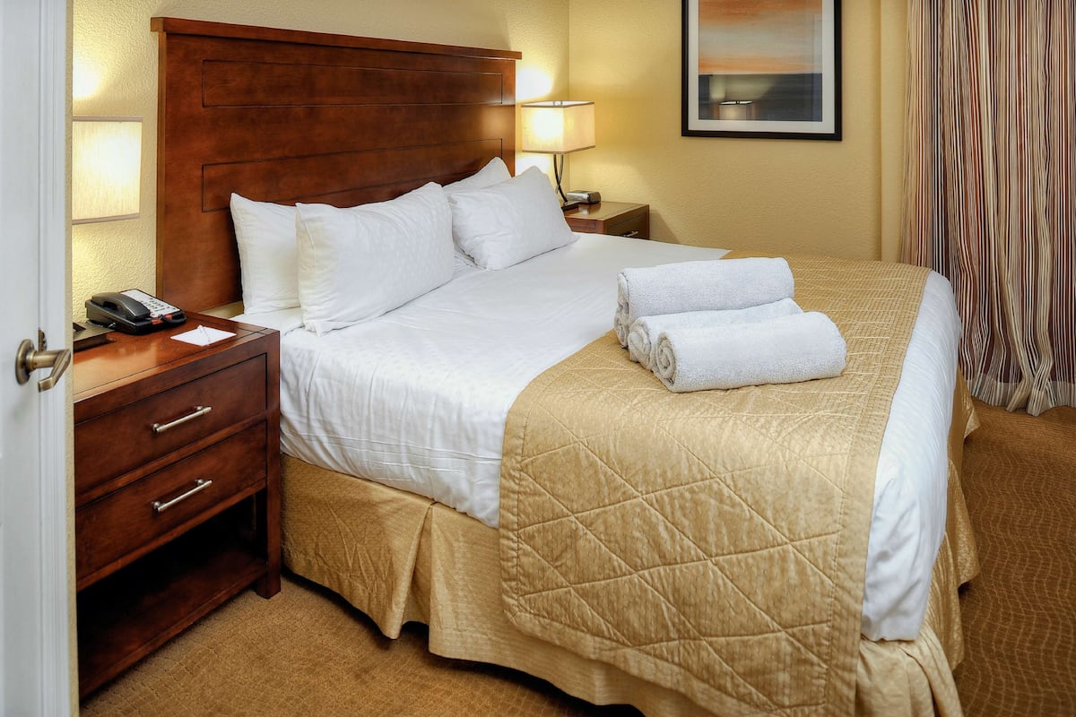Quarto de hotel com cabeceira de madeira, cama com roupa de cama branca e colcha bege, luminária de cabeceira, telefone e mesa de cabeceira.