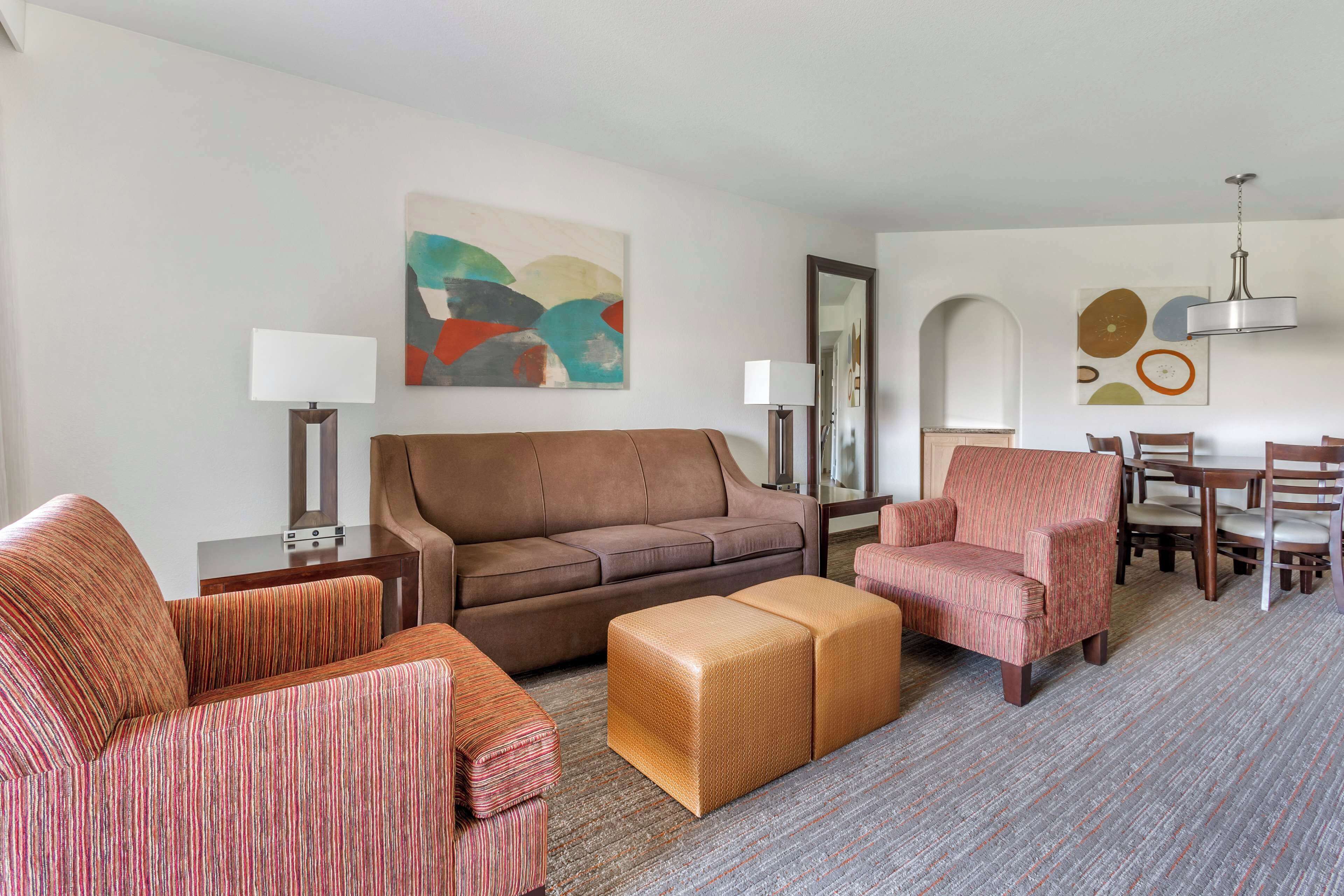 A modern living room with a brown sofa, striped armchairs, a woven ottoman, and a dining area with chairs and a table.