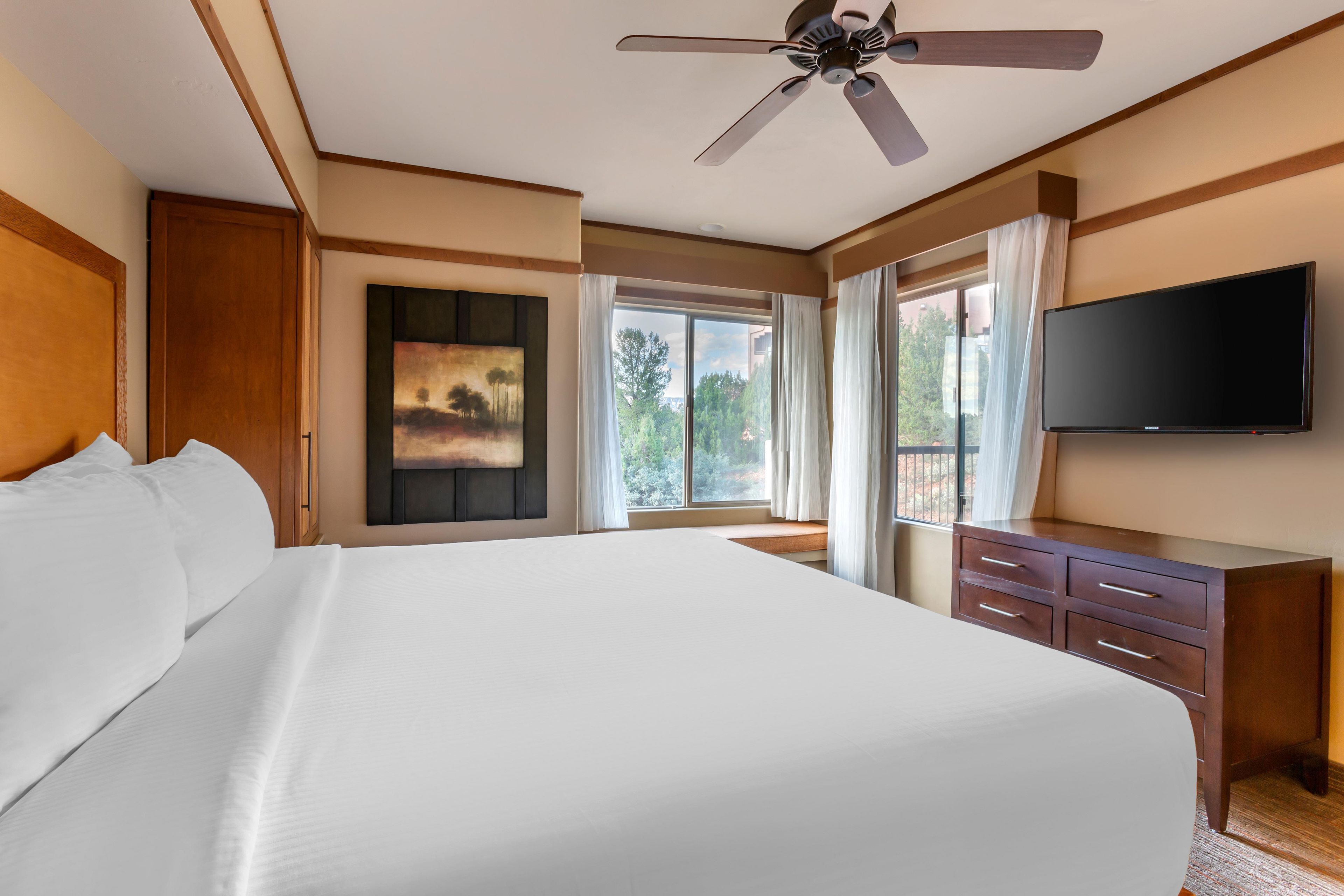Quarto de hotel com uma cama grande, ventilador de teto, televisão de tela plana, um quadro e uma janela com cortinas.