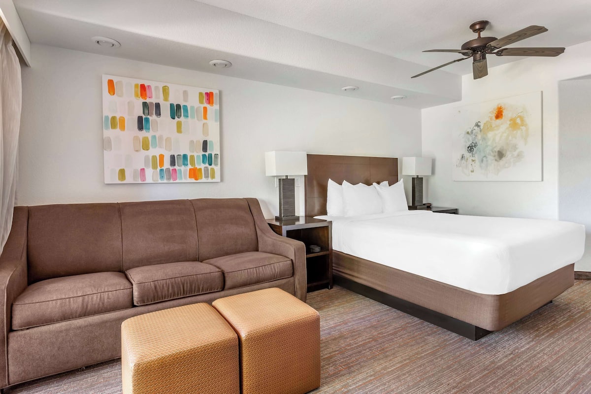 Quarto de hotel com sofá marrom, cama, um pequeno banquinho e duas luminárias fixadas na parede, além de um ventilador de teto.